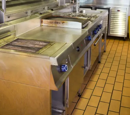 Interior de una cocina industrial con un mueble al centro que tiene un mueble con  estufa y tarja con repisas inferiores con puertas corredizas de acero inoxidable