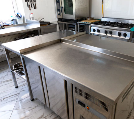Conjunto de tres mesas de acero inoxidable al centro de una cocina de restaurante, con una estufa y horno industrial al fondo