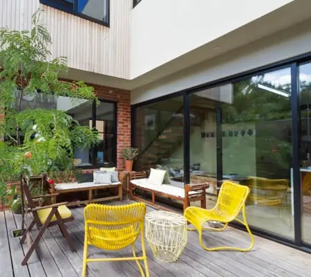 Vista lateral del patio de una casa con muebles para exterior y un árbol mediano del lado izquierdo, al fondo se ve una amplia puerta junto al resto de la casa de dos plantas