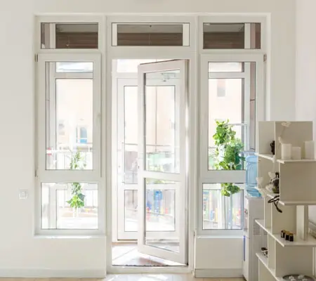 Vista frontal de una ventana color blanco y vidrio, se encuentra en un cuarto que da a un balcón, del lado derecho hay un mueble de madera con varias repisas.