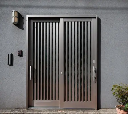 Vista frontal de una puerta de aluminio corrediza color gris metálico ubicada al frente de una pequeña casa color gris, a la derecha hay pequeñas macetas decorativas